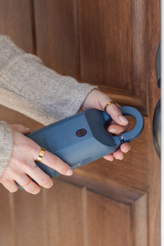 A person's hands wearing gold rings and a light textured sleeve hold a blue Supra electronic lockbox near the dark doorknob of a wooden door.