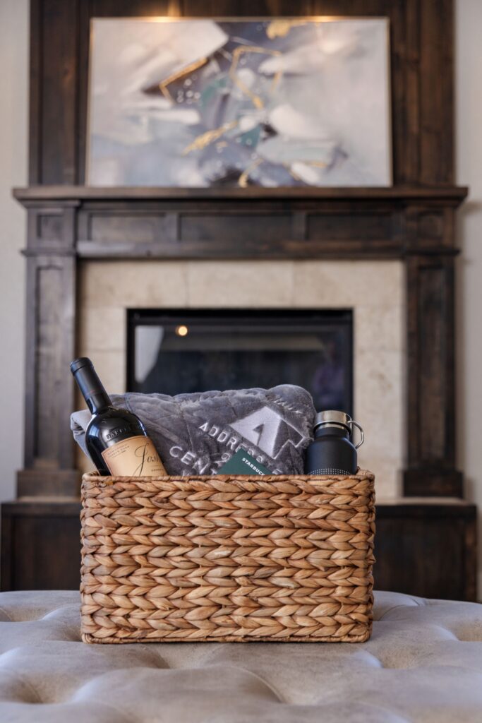 A woven gift basket containing a bottle of wine, a grey blanket, and a black water bottle sits on a tufted ottoman in front of a fireplace.