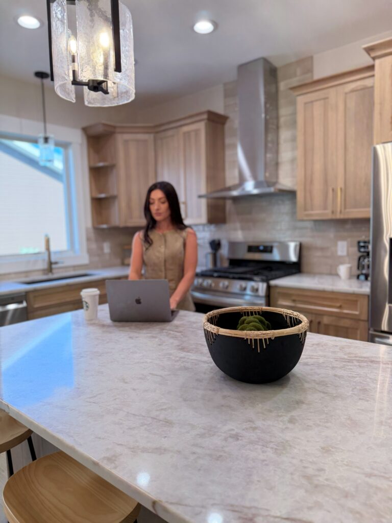 A woman in a beige top works on a laptop at a kitchen island, with a black bowl in the foreground and light wood cabinets and stainless steel appliances in the background.